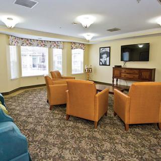 A cozy living room with upholstered chairs facing a television, featuring large windows and patterned carpet.
