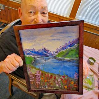 An elderly artist proudly displays their vibrant landscape painting featuring mountains and a serene lake.