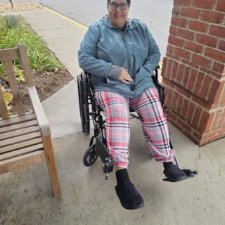 A woman sitting in a wheelchair outdoors, dressed in comfortable plaid pants and a casual jacket.