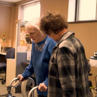An elderly woman receives assistance while using a walking aid, with a caregiver providing support.