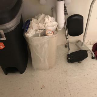 An indoor scene showing a trash can filled with garbage beside sink pipes.
