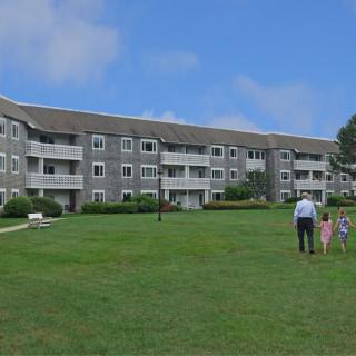 A group of people walking towards a large residential building on a grassy lawn under a blue sky.