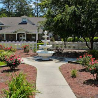 A beautifully landscaped garden with a central fountain, surrounded by flowers and trees, leading up to a quaint building.