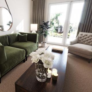 A modern living room with a green sofa, beige armchair, and a wooden coffee table adorned with flowers, illuminated by natural light.