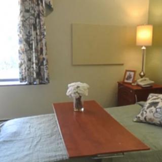 A hospital bedroom with a bedside table and lamp, featuring a vase of flowers and window curtains.