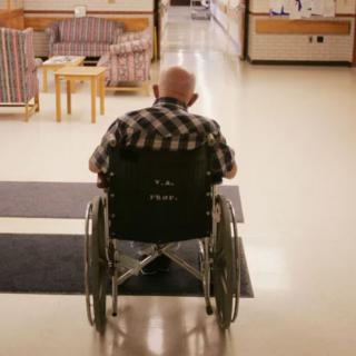An elderly person in a wheelchair in a nursing home corridor.