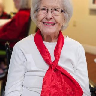 A cheerful elderly woman wearing a red scarf, sitting in a wheelchair with a warm smile.