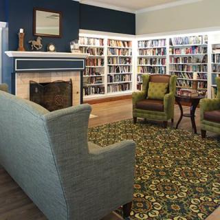 A cozy library with bookshelves, armchairs, and a sofa surrounding a fireplace.