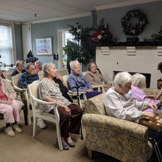 A group of elderly people seated in a living room setting, seemingly engaged in an activity.