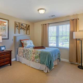A cozy bedroom featuring a single bed with floral bedding, a wooden nightstand, large window, and ambient lighting.