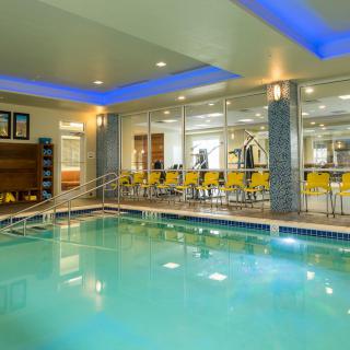 A modern indoor swimming pool with a seating area and glass walls leading to a fitness center.