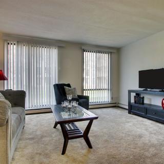 A cozy living room featuring a sofa, armchair, coffee table, and TV with large windows and vertical blinds.