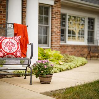 A cozy patio bench with a patterned pillow and potted plant against a brick house facade.