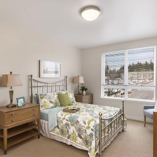A cozy bedroom with a metal frame bed, floral bedding, and wooden furniture, illuminated by natural light from a large window.