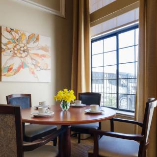 A neatly set dining table with chairs and a vase of flowers, next to a large window with golden curtains in a well-decorated interior.