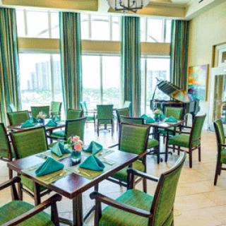 A brightly lit dining area with green chairs, neatly set tables, and a grand piano by large windows.