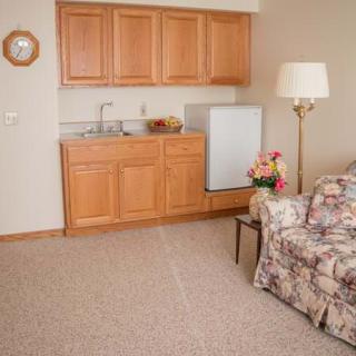 A small kitchenette with wooden cabinets and a mini fridge, accompanied by a floral sofa and floor lamp in a carpeted room.