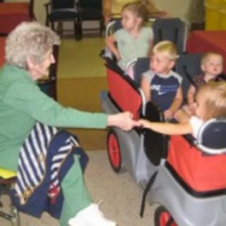 An elderly woman interacts with children seated in a stroller inside a daycare setting.