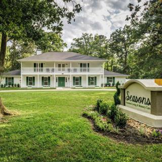 A colonial-style house surrounded by a lush green lawn and trees, with a sign in the foreground under a cloudy sky.