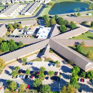 An aerial view of a building complex surrounded by trees and a pond, featuring a spacious parking lot.
