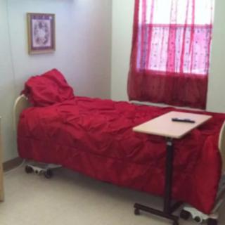 A simple room featuring a hospital bed with red bedding, a tray table, and a window with red curtains.