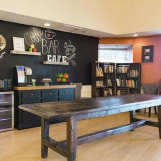 A modern cafe interior featuring a large wooden table, a chalkboard menu, and a cozy bookshelf.