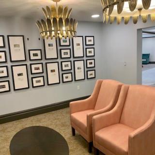 A modern waiting room featuring pink chairs, decorative chandeliers, and a gallery wall of framed artwork.