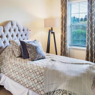 A cozy bedroom with a neatly made bed, featuring patterned bedding and pillows, next to a window with drapes and a floor lamp.