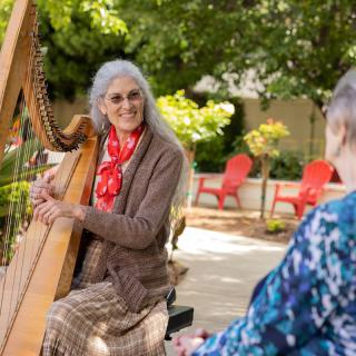 An elderly woman plays the harp in a garden while another person listens.