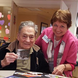 An elderly man and a woman smiling together indoors, with the man holding a photograph.