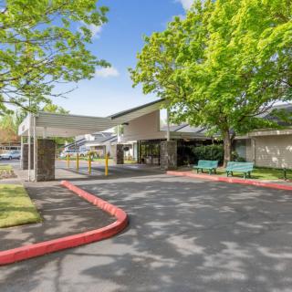 A sunny day view of the entrance to a building surrounded by trees and a parking lot.