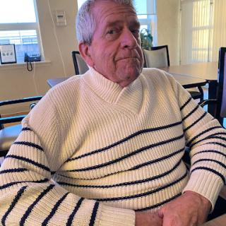 An elderly man in a striped sweater sitting in a cozy indoor setting.