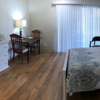 Bright bedroom with wooden floor, featuring a bed, desk, and window blinds.