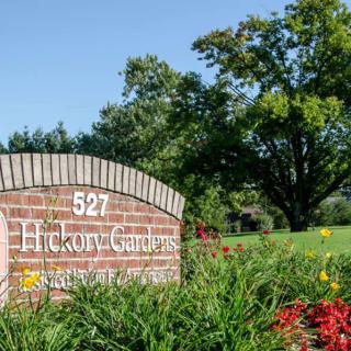 A brick sign with the name 'Hickory Gardens' surrounded by lush greenery and flowers.