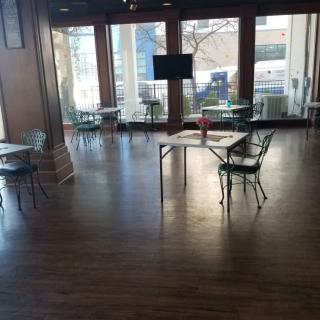 A bright, empty room with wooden floors, scattered tables and chairs, and large windows letting in natural light.
