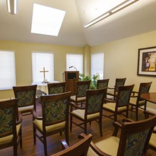 A small chapel with arranged chairs facing a pulpit and cross, illuminated by natural light from large windows and a skylight.