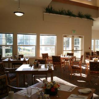 A sunlit dining area with neatly arranged tables and chairs, large windows providing a view outside.