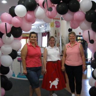 Three people standing under a colorful arch of pink, white, and black balloons, with one person wearing a poodle skirt.