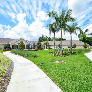 A scenic view of a house with a lush green lawn, palm trees, and a winding pathway under a partly cloudy sky.