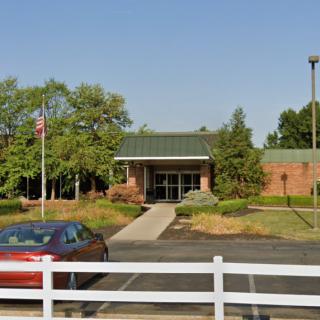 A small brick building with a green roof, surrounded by trees and a parking lot.
