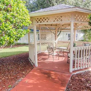 A charming garden gazebo with patio furniture, surrounded by lush greenery on a sunny day.