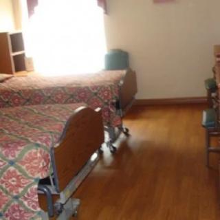 A hospital room with two beds and wooden flooring, illuminated by natural sunlight.