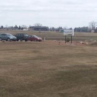A view of a parking lot with several cars and a basketball hoop in an open field under a cloudy sky.