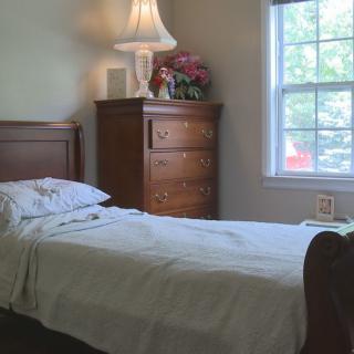 A cozy bedroom with a single bed, lamp, dresser, and window view.