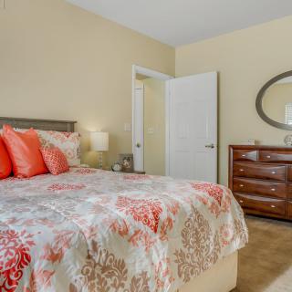 A cozy bedroom with a bed featuring floral bedding, a wooden dresser with an oval mirror, and soft lighting.