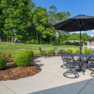 A sunny patio with outdoor furniture and an umbrella, surrounded by a landscaped garden.
