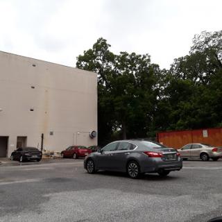 A gray car navigating a parking lot beside a tall building surrounded by trees.