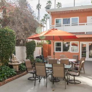 A cozy backyard patio with a table and chairs under an orange umbrella, surrounded by greenery.