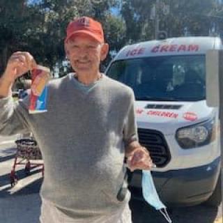 A man wearing a cap stands outdoors holding ice cream in front of an ice cream van on a sunny day.