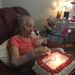 An elderly woman sitting in a chair prepares to blow out candles on her birthday cake.